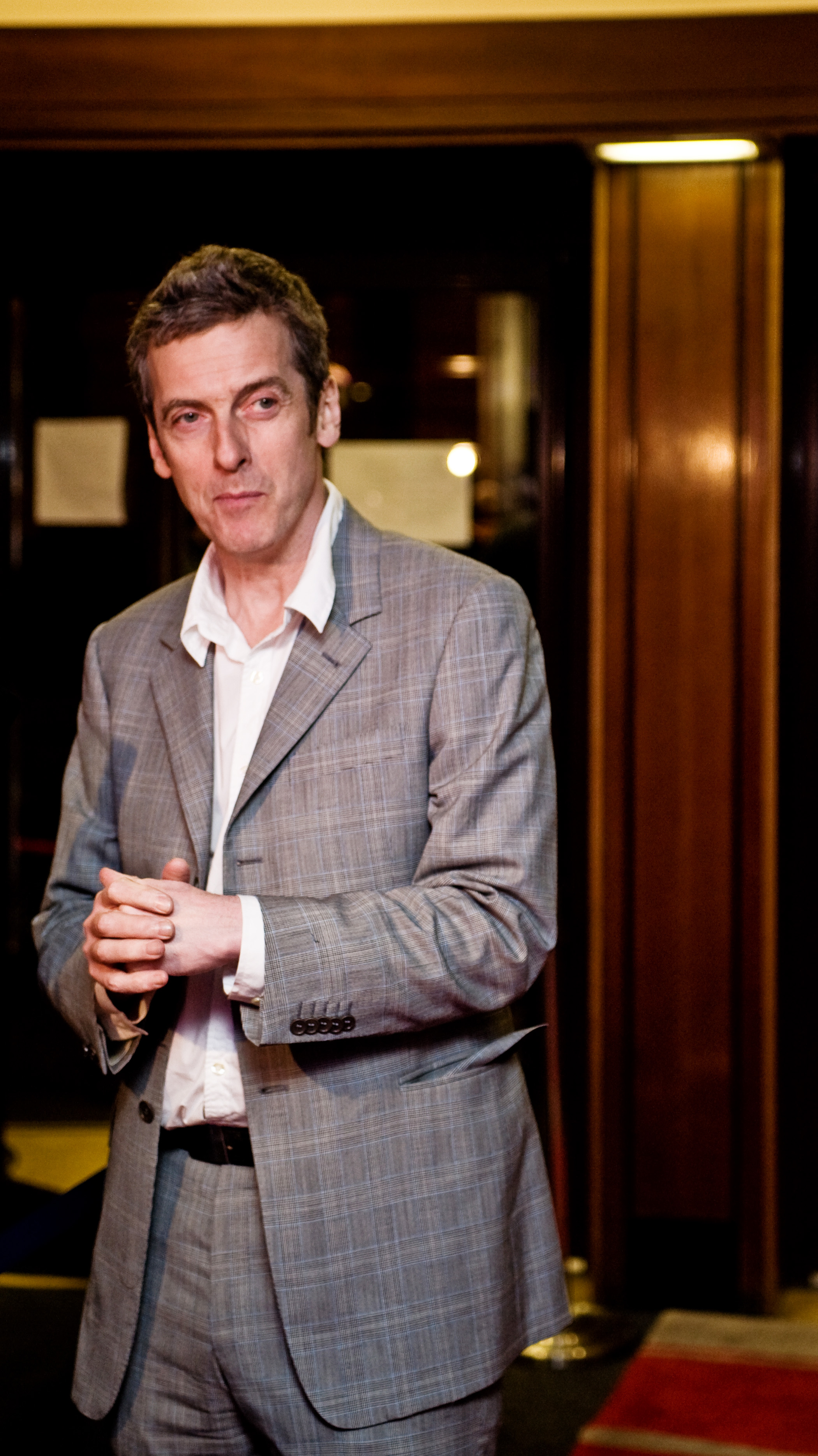 A photograph of Peter Capaldi at a showing of the film In The Loop. He is a man in his early 50s dressed smartly in a grey, hatched suit and white, collard shirt with no tie. He is looking to one side and clasping his hands in a slightly menacing way.